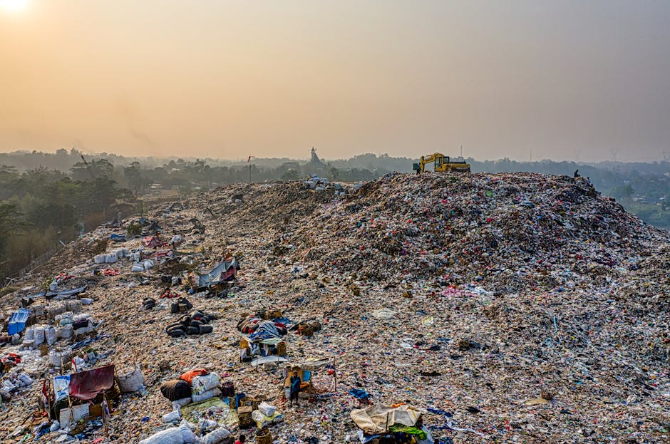 A large landfill site filled with a vast heap of mixed waste materials, including plastic bags, cardboard boxes, fabric items, tires, and miscellaneous household rubbish, all piled high under an overcast sky with a hazy atmosphere. The waste is spread across a sloping terrain, with several makeshift shelters constructed from tarps, cloth, and discarded materials positioned among the rubbish. A yellow excavator is visible at the top of the heap, indicating active waste management or landfilling processes. In the background, distant trees and a faint skyline are seen, with soft, diffused lighting from the cloudy sky. The scene illustrates the scale of waste accumulation, relevant to the context of waste disposal regulations and bulk waste management, as discussed in the webpage about Barnet rules for bulk waste and skip hire, by Man with Van Brent Cross.