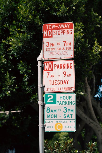 A vertical metal pole mounted with four parking restriction signs situated outdoors against a backdrop of green leafy trees. The top sign indicates a tow-away zone with no stopping allowed from 3 pm to 7 pm, except on Saturdays and Sundays, including street cleaning times. The second sign below specifies no parking from 7 am to 9 am on Tuesdays for street cleaning. The third sign features a teal background with the number '2' and states that parking is permitted for two hours between 8 am and 3 pm from Monday to Saturday, with an exception for vehicles with C permits. The bottom sign contains circular colour codes for parking permits. This signage is relevant for home relocations involving parking permits, loading, and moving logistics, as illustrated on the webpage for best parking permit tips for Barnet Council removals, provided by Man with Van Brent Cross.