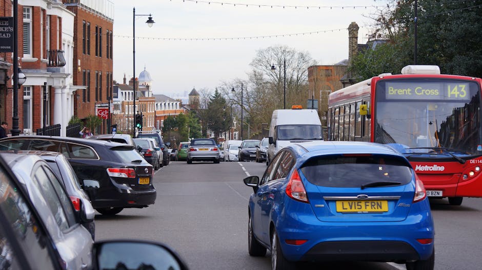 A busy street scene in Barnet, London, featuring multiple parked and moving vehicles, including a red Metroline bus with destination display showing 'Brent Cross 143', a blue hatchback with visible registration plate 'LV15 FRN', and several black and white cars lining both sides of the street. In the background, there are residential and commercial buildings, some with brick facades, street lamps, and trees with autumn foliage. A white van is approaching from the distance, likely used for home relocations or furniture transport, and street lighting is visible along the pavement. The scene depicts an active urban environment suitable for house removals and city-based moving services, with natural daylight illuminating the area and clear weather conditions.