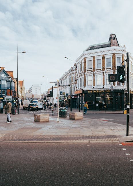 A busy urban street scene in Brent Cross featuring a corner building with a classic architectural design, including white decorative trim and large windows on the upper floors, and a pub named 'The World's End' situated on the ground floor. The street is active with pedestrians walking on the sidewalks, some crossing the road at a pedestrian crossing with a green signal, and vehicles including cars and a bus moving along the road. In the foreground, there are several wooden crates placed on the paved pavement, which appears to be part of a home relocation or moving process. The scene is captured during daytime with partly cloudy skies, and street furniture such as lampposts and traffic lights are visible, along with some overhead tram or trolleybus wires. The image is relevant to house removals and furniture transport, illustrating the logistical environment surrounding property moves, as seen through the exterior of a typical NW4 West London street, supported by [COMPANY_NAME] in providing professional removal services.