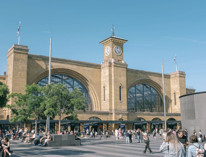 A large, historic train station building made of light-colored brick features two tall, arched glass windows and a central clock tower with a clock face on each side, set against a clear blue sky. Flagpoles with Union Jack flags are positioned on both sides of the building. In the foreground, a busy outdoor area is populated with people sitting on benches, walking, and standing, some carrying bags or backpacks. Several trees with green foliage are situated in front of the station, partially obscuring the lower section. The scene includes outdoor lighting fixtures and a paved plaza where man with Van Brent Cross may conduct home relocation or furniture transport activities, reflecting a vibrant urban environment suitable for house removals and moving services.