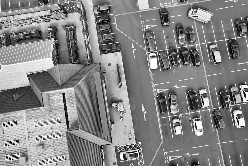 An aerial view of a large parking lot adjacent to a building with a sloped roof, seen from above. The parking area contains numerous cars parked in marked spaces, with some driving through lanes and others waiting to be parked. The lot is partially filled, with empty spaces available. There are several vehicles, including a van, parked near the building’s entrance, which is connected to a paved walkway. The building features a combination of roof styles, including pitched roofs and flat sections, with visible roofing materials such as tiles and metal sheeting. Nearby, there are cleaning supplies and cardboard boxes positioned on the pavement close to the building, suggesting ongoing activity related to home relocation or moving preparations. This setting resembles an area where professional removals services, such as those provided by Man with Van Brent Cross, might organize loading and unloading operations during furniture transport and house moving processes. Bright daylight illuminates the scene, highlighting the organized arrangement of vehicles and the environment for a home or business move.