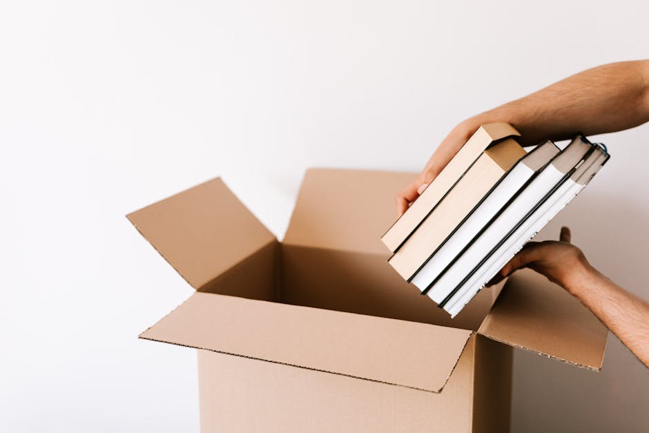 A person placing a stack of five books into a partially open cardboard box, with the person's hands visible holding the books and supporting the box. The books have beige, black, and white covers and are being packed carefully, suggesting a process of packing and moving items during a house relocation. The background is plain white, emphasizing the box and books. The image relates to packing and furniture transport services offered by Man with Van Brent Cross in the context of home removals and logistical preparations for a smooth move, as referenced on the page about bulky item moves on Clitterhouse Road.