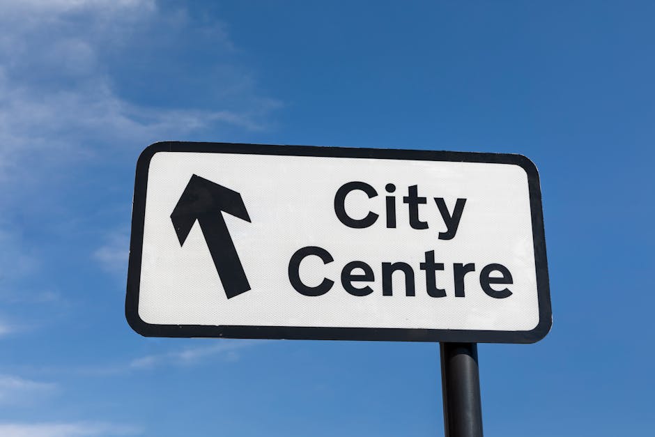 A rectangular road sign mounted on a black pole against a clear blue sky, indicating directions with a black arrow pointing straight ahead. The sign displays the words 'City Centre' in bold black lett