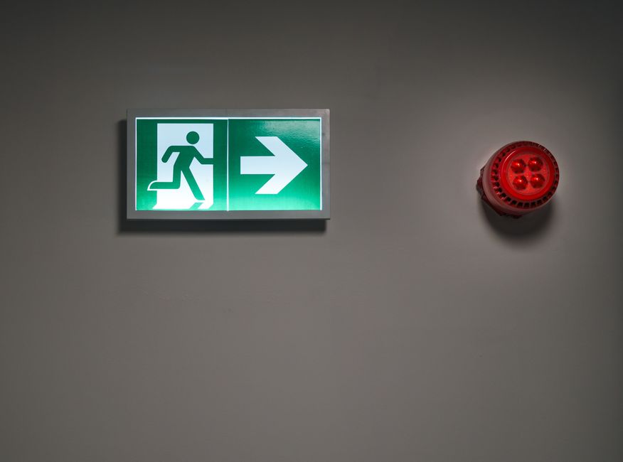 A green emergency exit sign with a pictogram of a person walking up stairs and an arrow pointing to the right is mounted on a plain grey interior wall. To the right of the sign, there is a red circular fire alarm or warning device attached to the wall. The lighting is even, with no visible window or natural light sources, indicating an indoor setting that could be part of a commercial or residential property undergoing a house relocation process. The sign and alarm are positioned at eye level, with the smooth wall surface providing clarity for quick identification during emergency evacuations, supporting safe and efficient house removals or emergency evacuation moves conducted by Man with Van Brent Cross.