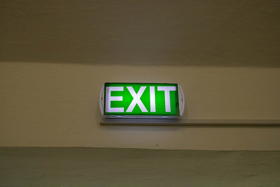 A green and white illuminated emergency exit sign mounted on a beige indoor wall, positioned above a horizontal metal rail or conduit. The sign displays the word 'EXIT' in bold capital letters, indicating an evacuation route. The ceiling above the sign is neutral-toned and plain, with soft lighting illuminating the sign clearly for visibility. This image is taken inside a building, likely near a corridor or stairwell, and may be part of a context involving emergency evacuation planning or building safety protocols. No moving equipment, furniture, or packaging materials are visible; the focus is solely on the illuminated exit sign, which is relevant to the concept of safe building exits in house or building removals and emergency protocols, aligning with services provided by Man with Van Brent Cross.
