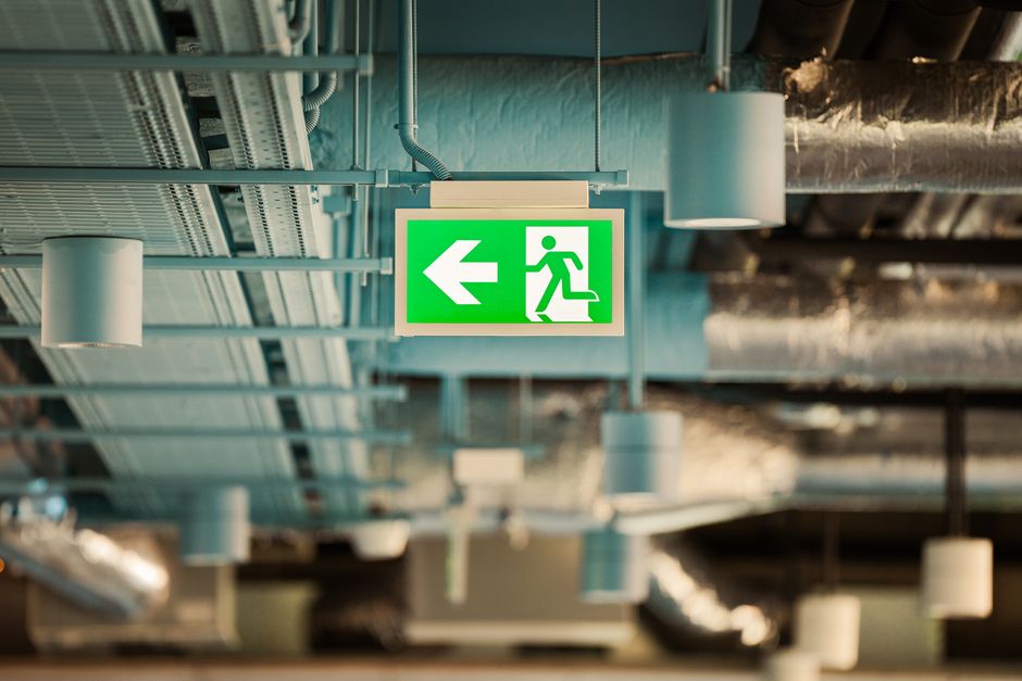 A green emergency exit sign with a pictogram of a person walking up stairs and an arrow pointing to the right is mounted on a plain grey interior wall. To the right of the sign, there is a red circular fire alarm or warning device attached to the wall. The lighting is even, with no visible window or natural light sources, indicating an indoor setting that could be part of a commercial or residential property undergoing a house relocation process. The sign and alarm are positioned at eye level, with the smooth wall surface providing clarity for quick identification during emergency evacuations, supporting safe and efficient house removals or emergency evacuation moves conducted by Man with Van Brent Cross.