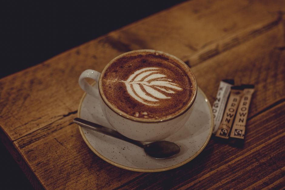 The image shows a disposable coffee cup with a white plastic lid, featuring the Costa Coffee logo and branding in white lettering on a ribbed, dark red paper sleeve. The cup is placed on a table or co