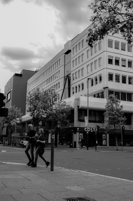 A black and white photograph showing a busy urban street scene outside a modern multi-storey office building with large windows and a white facade. In the foreground, two movers from Man with Van Brent Cross are walking across the pavement, dressed in casual clothing. One is carrying a cardboard box, while the other appears to be balancing a small piece of furniture wrapped in protective padding. The pavement is lined with young trees and street furniture, including a lamppost and a bus stop sign. Parked vans and a COTY store are visible along the street, with some vehicles partially obscured by the movers. The cloudy sky overhead casts a diffuse light over the scene, highlighting the ongoing logistics involved in home relocation or furniture transport, typical of house removals and packing and moving services in the Brent Cross area.
