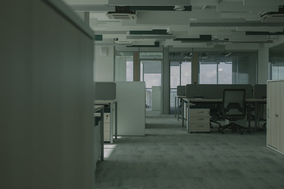 Inside a modern office space during a relocation process, with rows of work desks separated by low grey partitions, ergonomic swivel chairs, and small drawers under each desk. Clear plastic-wrapped cardboard boxes are placed on the desks and on the floor, with some being carried by movers using trolleys. The scene is illuminated by natural light coming through large glass windows and doors at the far end, revealing a balcony or outdoor area. The ceiling features a suspended panel design with integrated lighting and air conditioning vents. In the foreground, a partially visible moving blanket or padding covers a section of furniture or equipment being prepared for transport. The environment is tidy, and the setting reflects a professional office relocation, coordinated by Man with Van Brent Cross, involving careful packing, furniture transport, and loading processes for minimal business downtime.