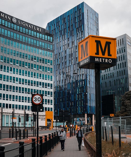 A street scene in front of modern office buildings featuring a prominent orange and black metro station sign with the letter 'M', indicating the Brent Cross area. The background shows multi-storey office blocks with glass and steel facades, including one with a 'MAN Signature' sign. The pavement is populated with pedestrians, some carrying bags, walking past black safety barriers and a 30 mph speed limit sign. Nearby, a small yellow kiosk or box is positioned close to the station entrance. The scene is captured during daytime with natural light, and the area appears busy and well-maintained, reflective of a central business district. This setting may relate to the logistical movement involved in house or office relocations, such as moving furniture or equipment through secure access points around Brent Cross.