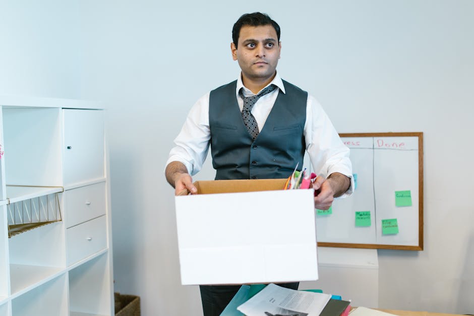 Inside a modern office space during a relocation process, with rows of work desks separated by low grey partitions, ergonomic swivel chairs, and small drawers under each desk. Clear plastic-wrapped cardboard boxes are placed on the desks and on the floor, with some being carried by movers using trolleys. The scene is illuminated by natural light coming through large glass windows and doors at the far end, revealing a balcony or outdoor area. The ceiling features a suspended panel design with integrated lighting and air conditioning vents. In the foreground, a partially visible moving blanket or padding covers a section of furniture or equipment being prepared for transport. The environment is tidy, and the setting reflects a professional office relocation, coordinated by Man with Van Brent Cross, involving careful packing, furniture transport, and loading processes for minimal business downtime.