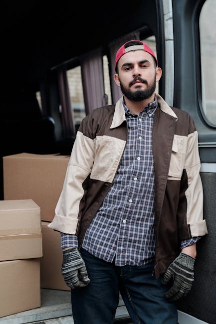 A male mover with a beard and wearing a red cap, brown and beige work jacket over a blue checkered shirt, and grey work gloves, standing inside a van loaded with cardboard boxes. The boxes vary in size and are made of brown corrugated cardboard, arranged along the side of the vehicle's interior, which has a dark lining. The man appears to be overseeing the home relocation process, with some boxes placed on the floor of the van and others stacked behind him. Natural lighting illuminates the scene, highlighting the details of the moving equipment and materials ready for furniture transport and packing during a same-day house removal service in Brent Cross. The image conveys professionalism and efficiency in the logistics of packing and loading items for a residential move, as provided by Man with Van Brent Cross.