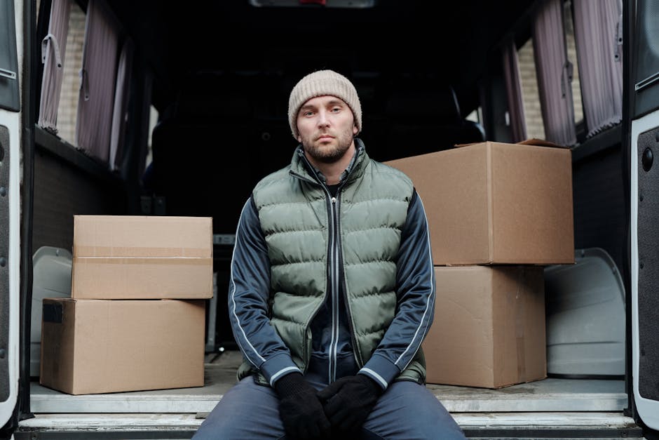 A male mover with a beard and wearing a red cap, brown and beige work jacket over a blue checkered shirt, and grey work gloves, standing inside a van loaded with cardboard boxes. The boxes vary in size and are made of brown corrugated cardboard, arranged along the side of the vehicle's interior, which has a dark lining. The man appears to be overseeing the home relocation process, with some boxes placed on the floor of the van and others stacked behind him. Natural lighting illuminates the scene, highlighting the details of the moving equipment and materials ready for furniture transport and packing during a same-day house removal service in Brent Cross. The image conveys professionalism and efficiency in the logistics of packing and loading items for a residential move, as provided by Man with Van Brent Cross.