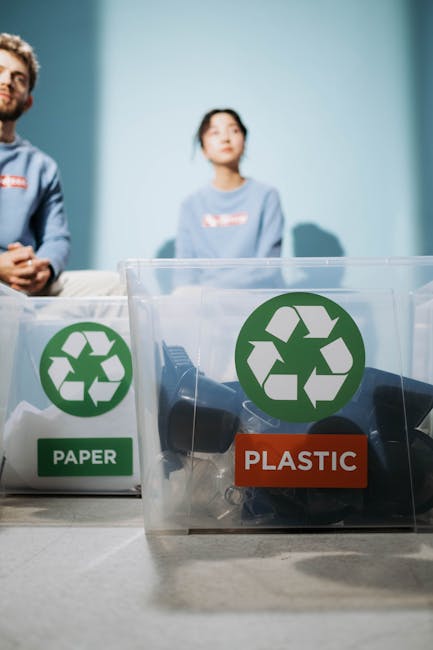 A person wearing a white shirt is holding a small piece of cardboard over a large blue recycling bin with a white recycling symbol on the front, positioned on a table against a light green wall. Next 